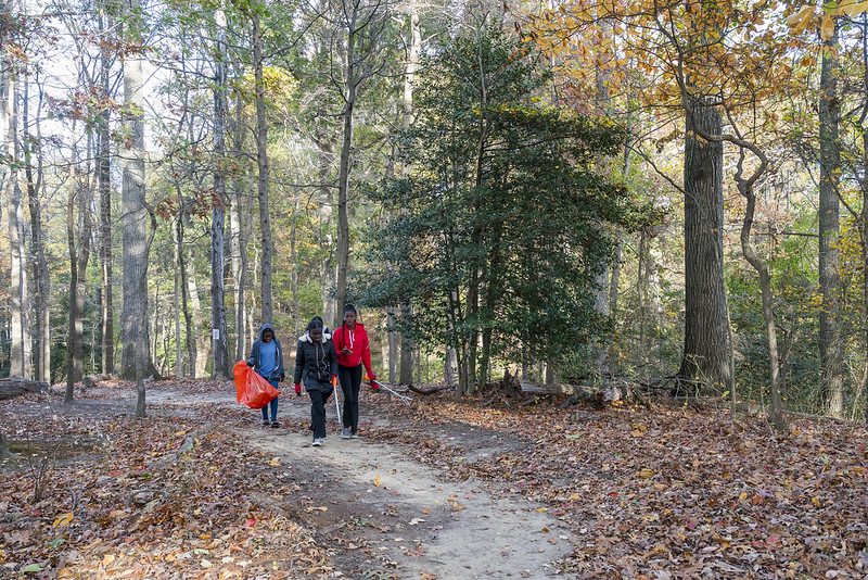 Three student volunteers walking down a gravel trail with trash bags in autumn. Trees, plants, and trees in the background.