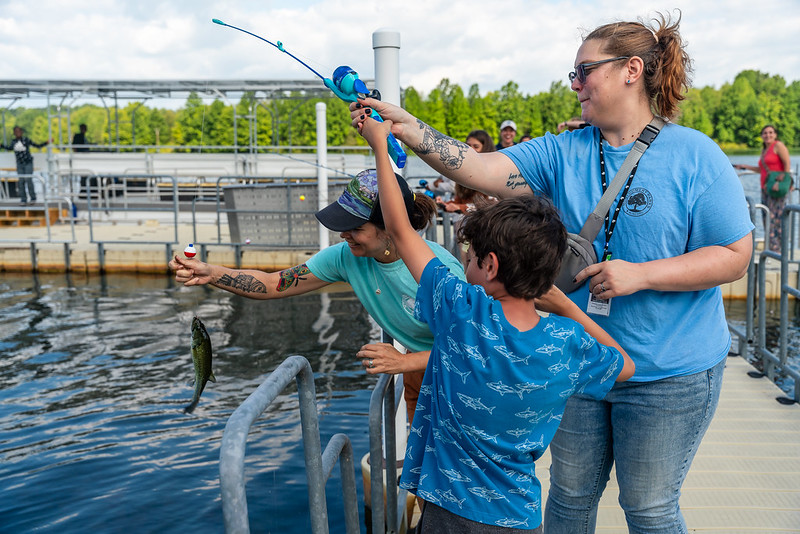 Program Access inclusion support staff assisting program participant with reeling in a fish caught in Little Seneca Creek and Park Naturalist pulling in the finish line of the fishing pole.