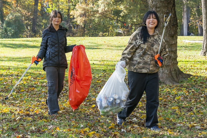Two student volunteers cleaning up trash from park. They carry trash bags full of trash. Green grass and tree in the background