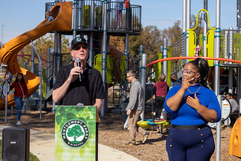 Sign language interpreter providing American Sign Language interpretation for speaker during a park ribbon cutting ceremony in front of the new playground.