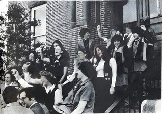 A black and white photo showing several teenage girls and young women standing on steps. Many raise their right fists in the air and have their mouths open.
