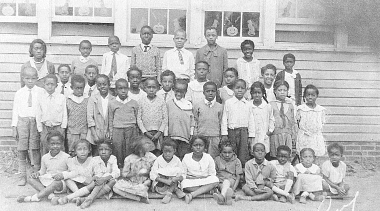 Black and white photo showing seated and standing African American children in front of a schoolhouse.