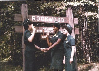 Three girls in Girl Scout uniforms stand in front of a sign reading "Rockwood." Two are holding hands and have their right hands raised as if to take a pledge.