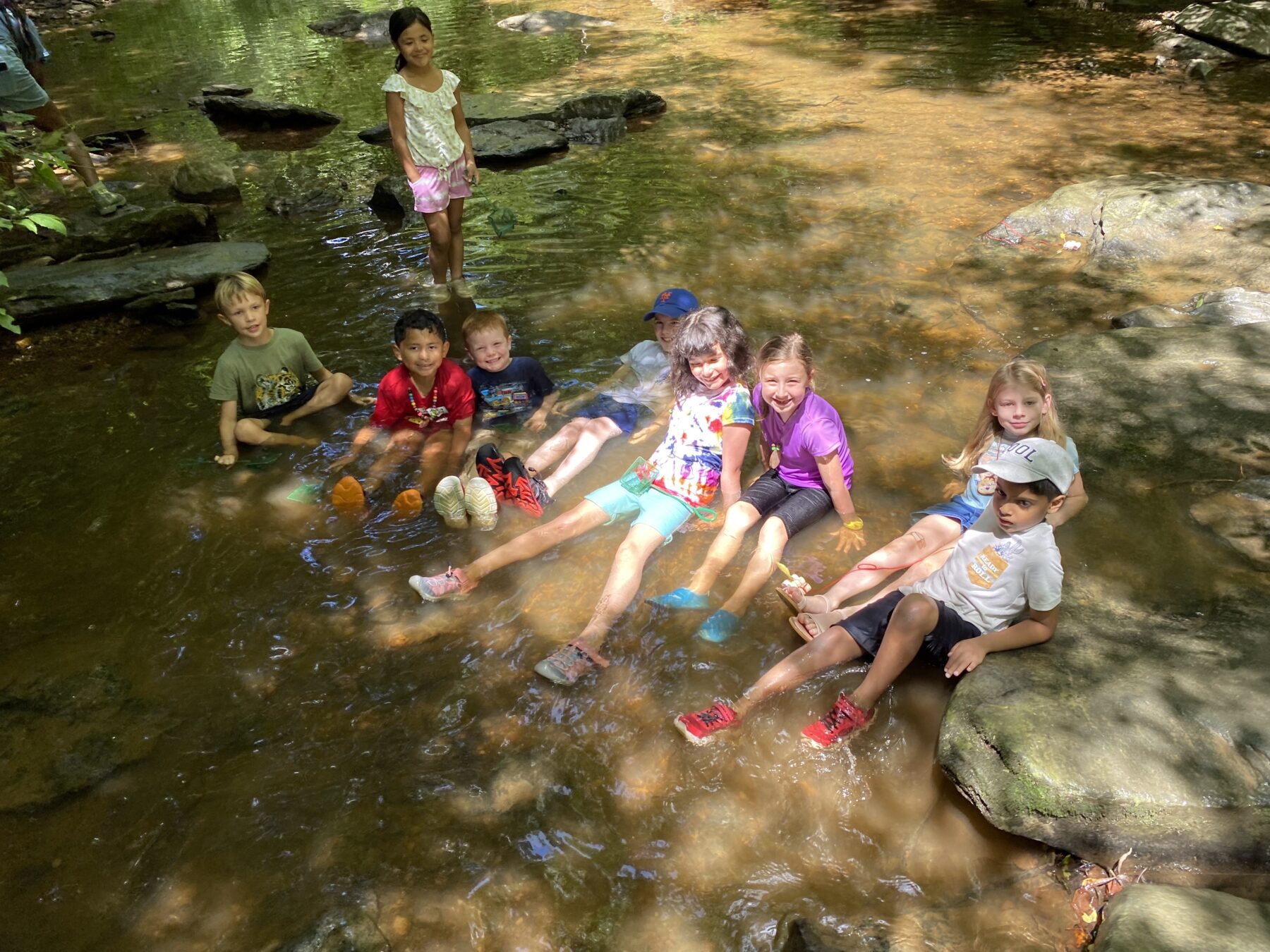 Nine kids are all sitting in a creek. The water is brown and the kids are dressed in shorts and t-shirts.