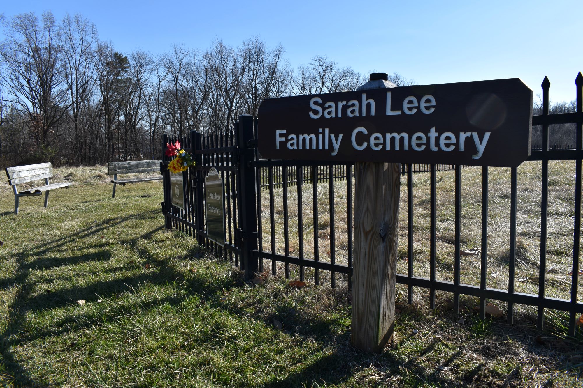A wooden sign reads "Sarah Lee Family Cemetery" posted outside of a metal fence surrounding a grassy area. Two benches and a stand of trees are in the background.