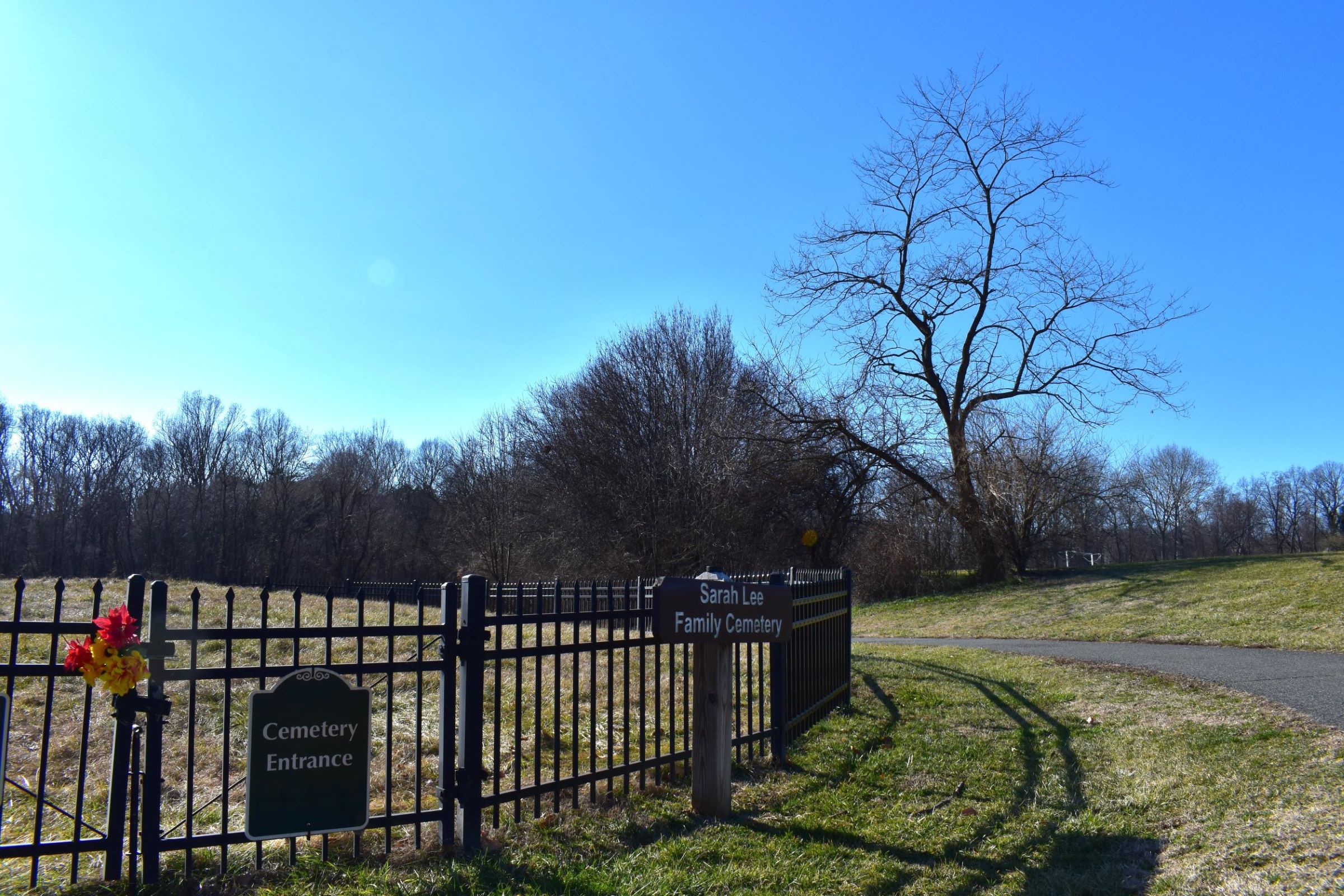 A metal fence surrounds a grassy area with a wooden sign posted in front that reads "Sarah Lee Family Cemetery." A walking path and stand of trees are to the right.
