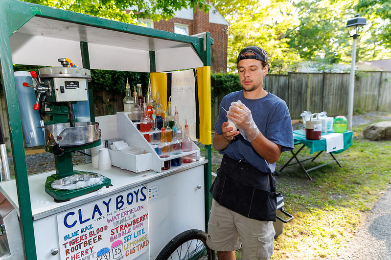 Clayboys shave ice staff member standing in front of cart with syrups and ice machine putting together a snowball