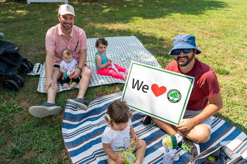 Tow parents with 3 young kids holding "We love Montgomery Parks" sign while sitting on picnic blankets 