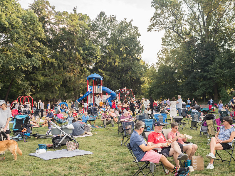 Large crowds sits on picnic blankets and chairs in large grassy field with kids on playground in the distance.