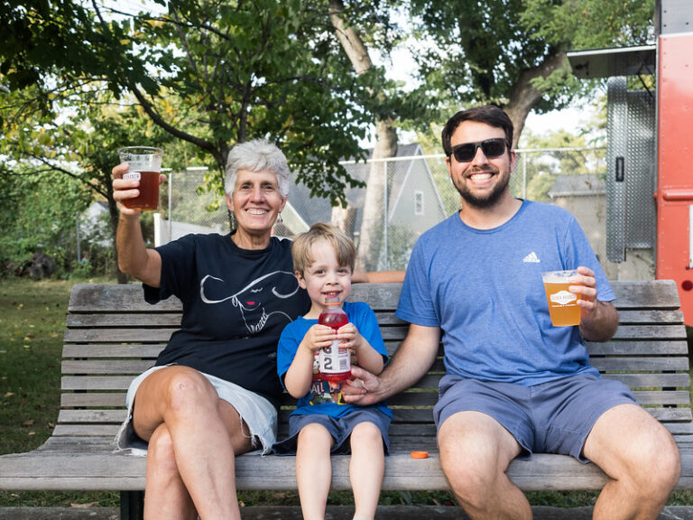 Family sits on bench. 2 adults are holding up beers while young boy sits between them drinking a bottle of Gatorade.