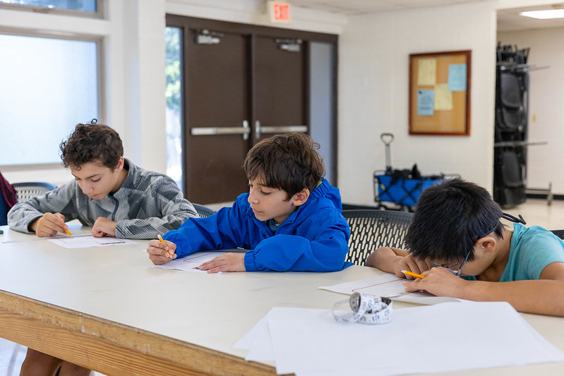 3 boys sit at a table, using pencils, paper, and rulers to design robots