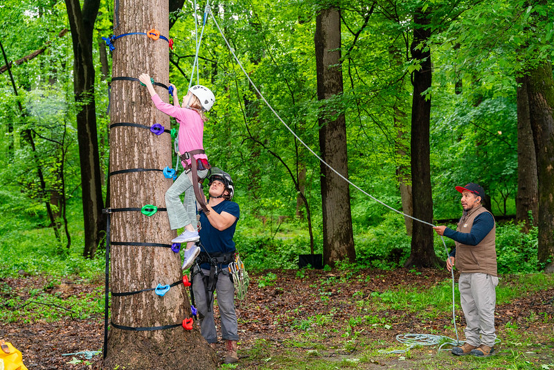 Child climbs a tree on a rope while staff helps her