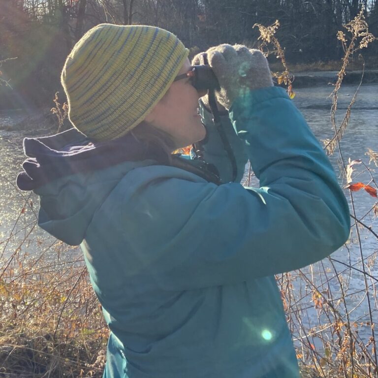 An adult, dress in blue winter clothing and a green knit hat, is looking ahead through a pair of binoculars. There is a frozen pond behind them.