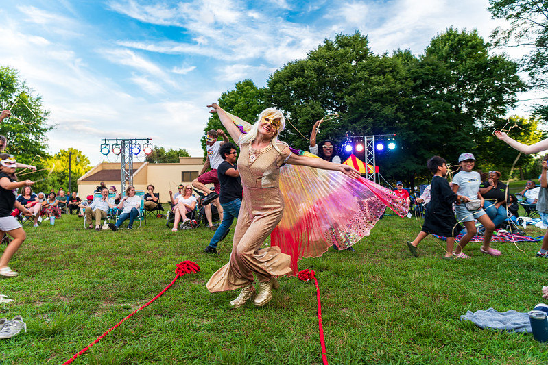 Performer dances with colorful wings while kids run around her with masks and strings