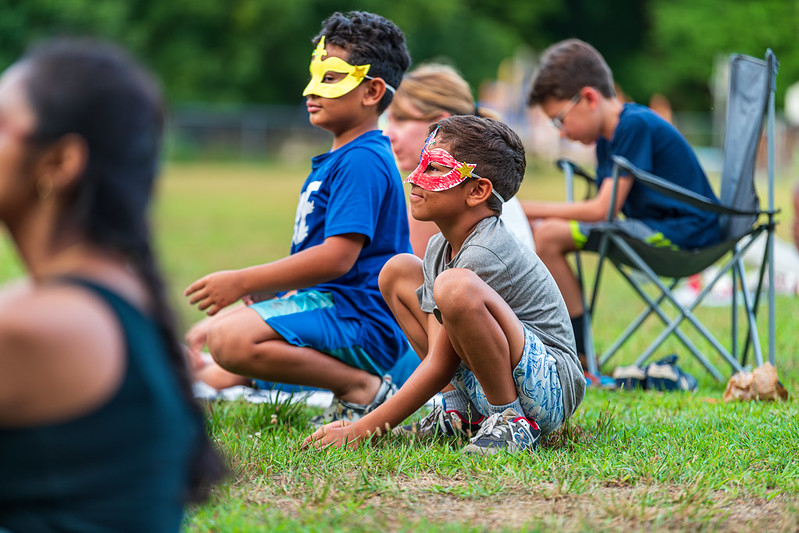 2 kids with masks sitting on the grass, watching the performance