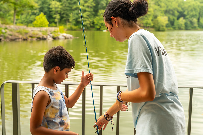 A child and a teen both hold the same fishing pole. They are standing by a fence and behind the fence is water that appears green in color.