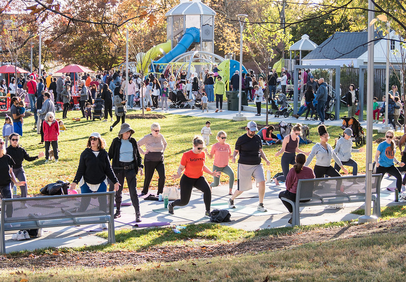 Photo at Caroline Freeland Urban Park showing people exercising in the lawn, kids playing on the playground and people gathering around the picnic tables, benches and swings in a sunny day.