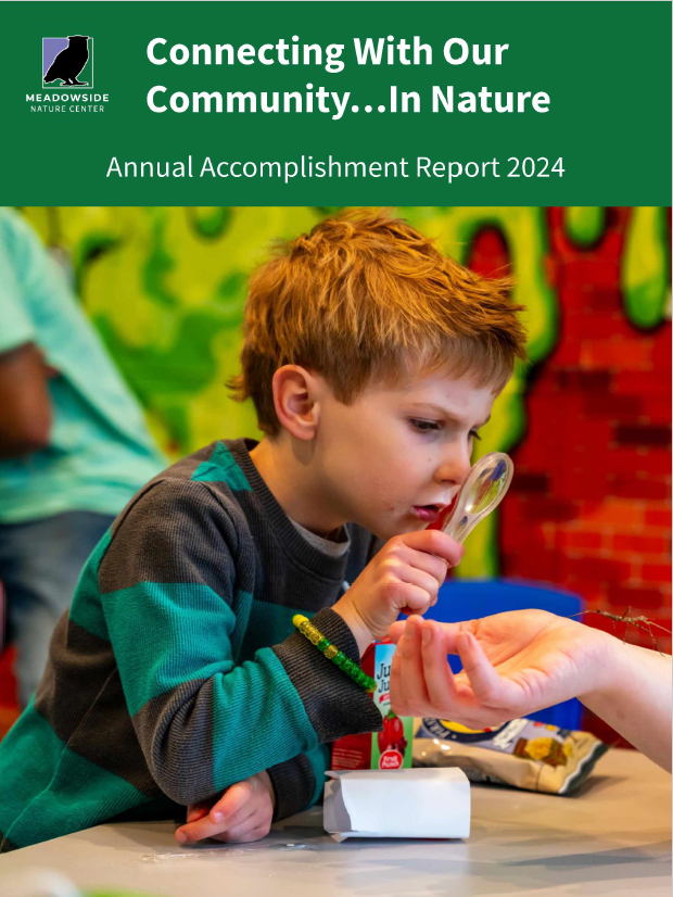 A close view of a boy using a magnifying glass to look at a stick bug that is walking on someone's bare arm. They are all inside a classroom. Text at the top of the image says " Connecting with our community in nature. Annual accomplishment report 2024."