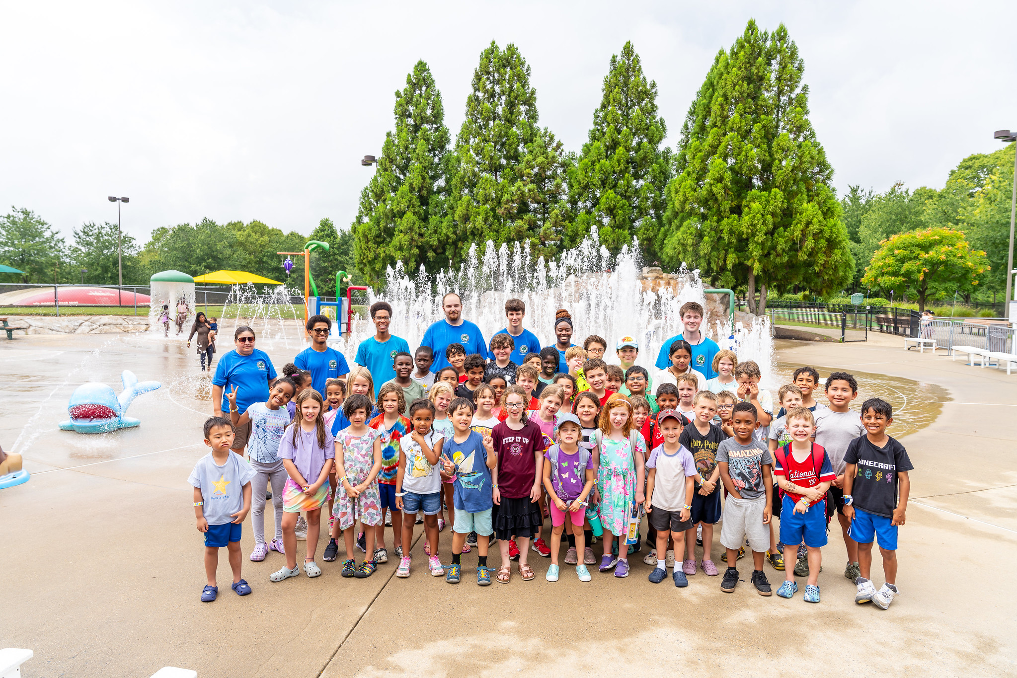 A group of campers and camp staff stand in front of water fountains at the splash park.