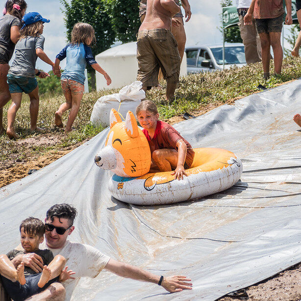 Children sliding down a big muddy hill using pool floats.