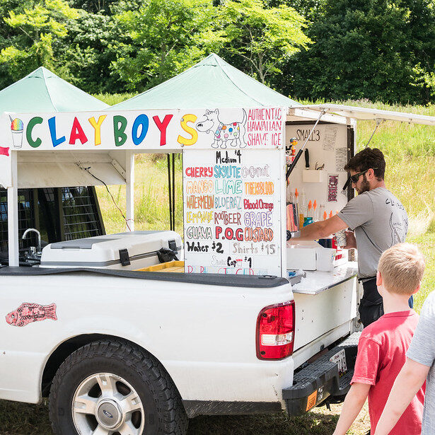 A person prepping shaved ice from a truck and two children waiting in line.