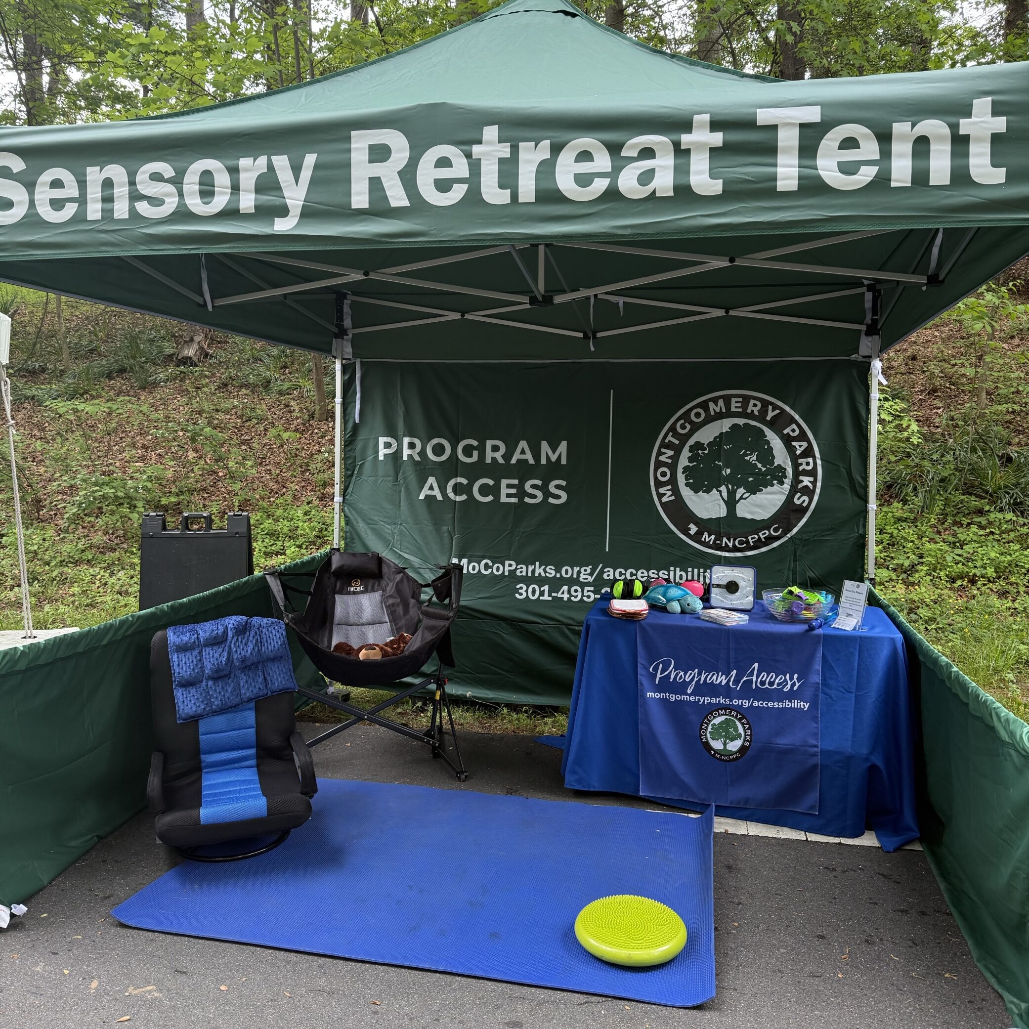 A green canopy tent with a banner that read "Program Access". Inside the tent are sensory seating and fidgets on top of a table.