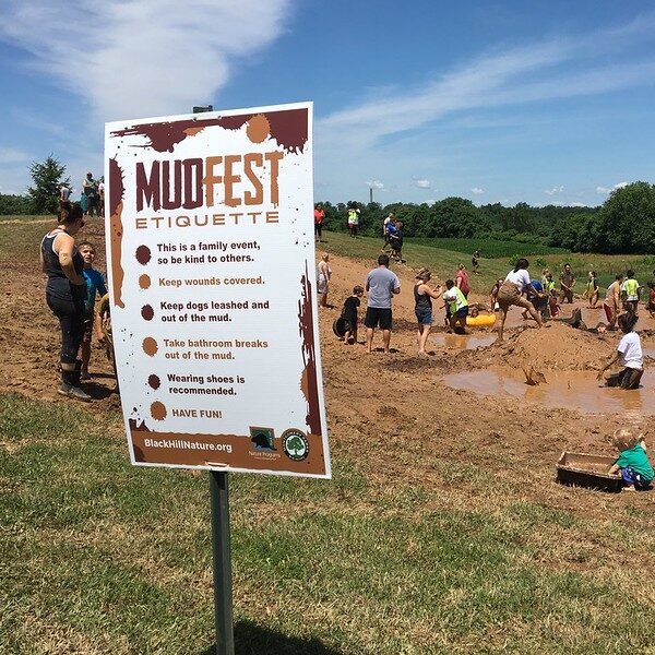 A "MudFest Mu Etiquette" sign with people playing in the mud in the background.