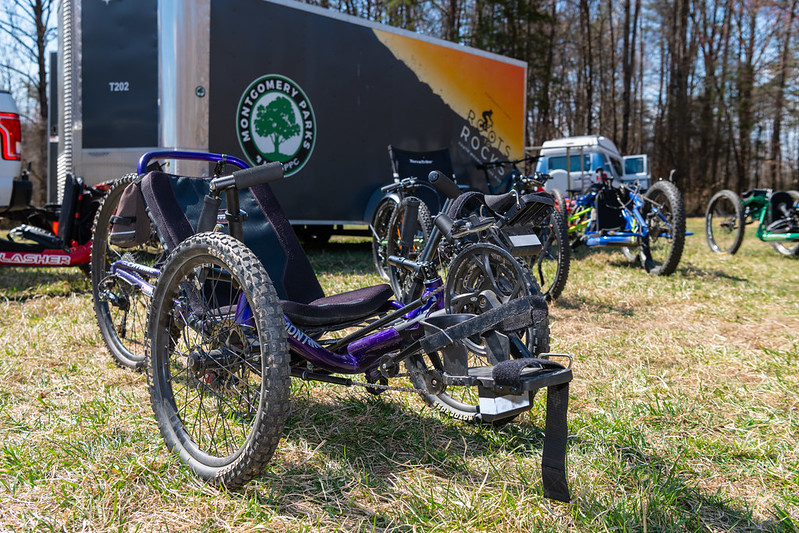 A hand-operated bike sitting on the grass in the foreground of other adaptive mountain bikes