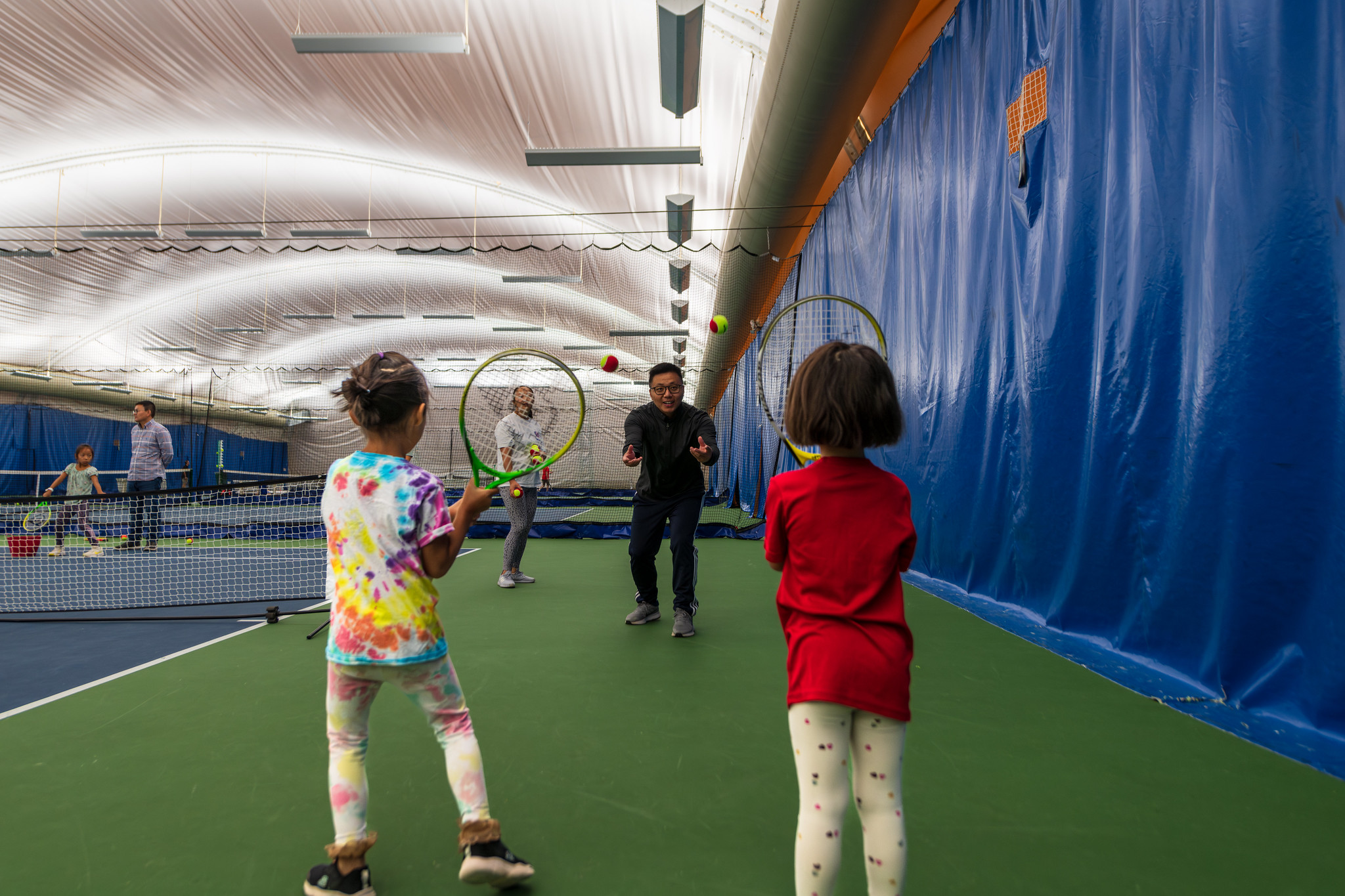 An adult throws a tennis ball toward two children holding racquets