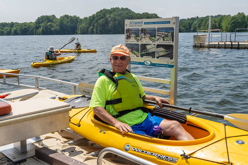 A person in an orange hat and green shirt and PFD using the Accessible boat launch system with a how-to- guide behind him.