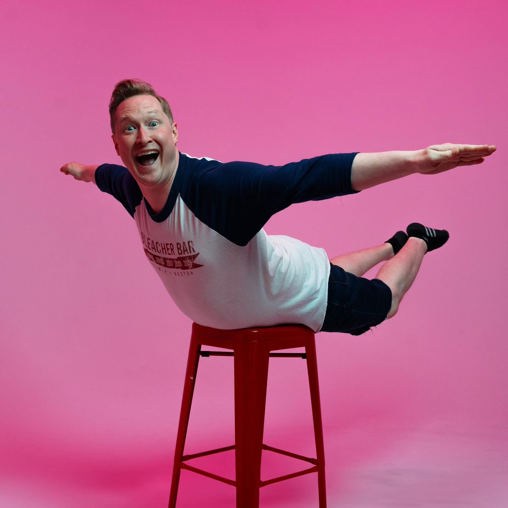 Comedian Brock Snyder poses on a stool laying on their stomach with their arms and legs out