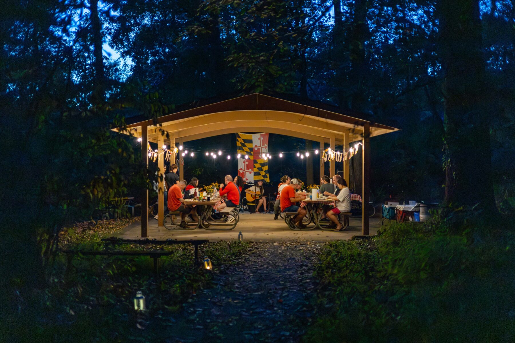 Several people are sitting at picnic tables outside under a pavilion. It is almost dark outside. The pavilion is light by white lights and there is a Maryland State flag haning in the background.