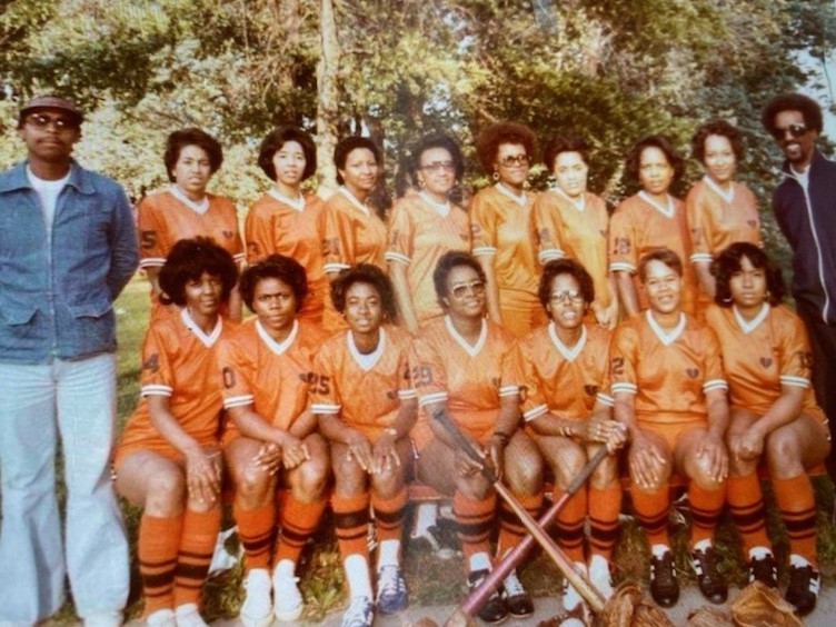 A color photo of the Heartbreakers, a women’s softball team from the mid-1970s. The women are wearing orange uniforms. Two male coaches stand on either side of the two rows of women, one row sitting and the other standing. They are outside in front of a small stand of trees.
