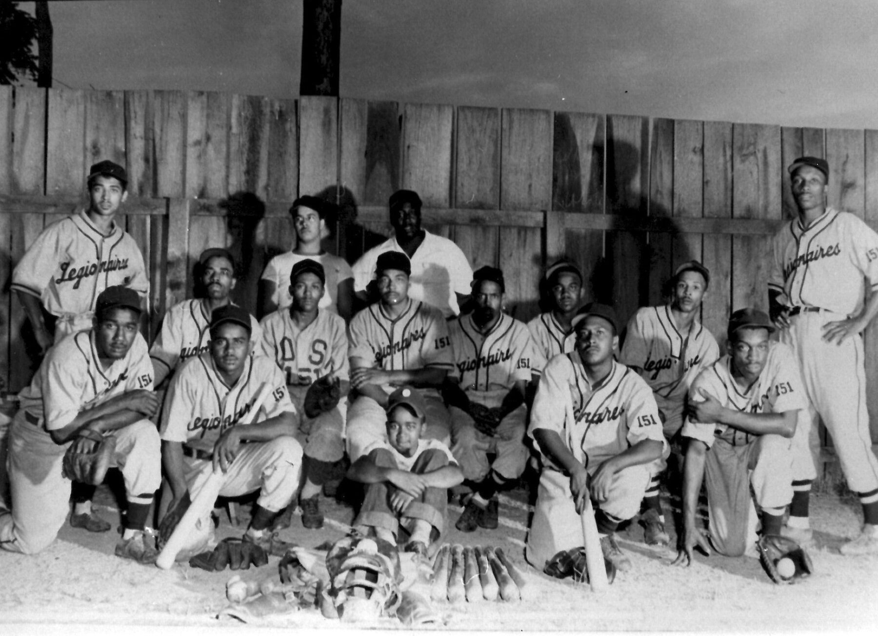 A black and white photo of Rockville’s American Legion 151 team, an African American men’s baseball team from the late 1940s. Two rows of men kneel or stand with bats, gloves, and baseballs in front of them. They are in front of an outfield fence and the photo was taken at night.
