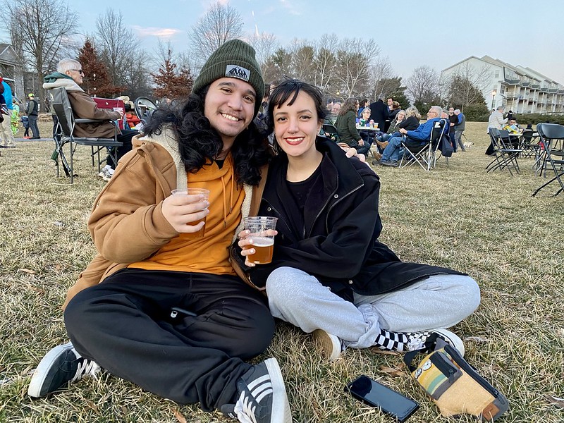 Young couple sits on lawn holding beers smiling for camera