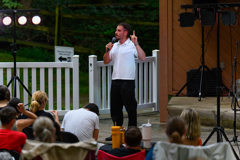 Comedian on stage with microphone holds one hand up