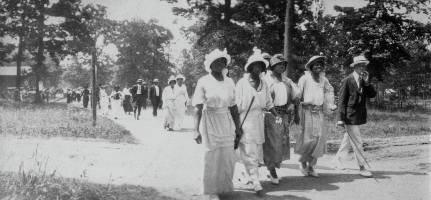A black and white photo showing a four African American women and one man walking along a dirt lane. There are more individuals in small groups walking in the same direction on the path behind them. All are dressed in suits and dresses.