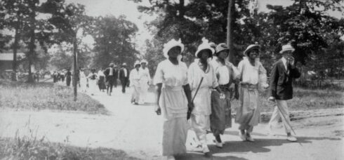 A black and white photo showing a four African American women and one man walking along a dirt lane. There are more individuals in small groups walking in the same direction on the path behind them. All are dressed in suits and dresses.