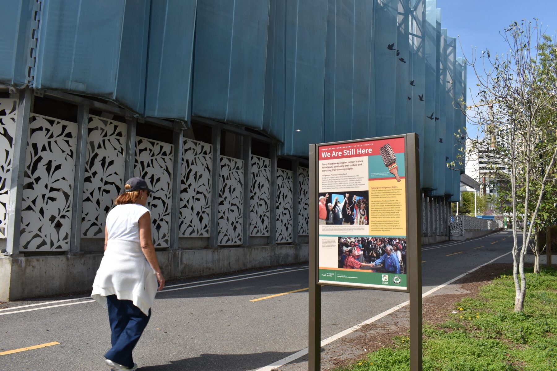 A woman walking on a paved trail past an interpretive sign titled "We Are Still Here."