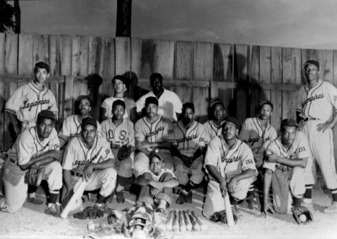 A black and white photo showing an African American men's baseball team posing for a team photo in front of a wooden outfield fence.