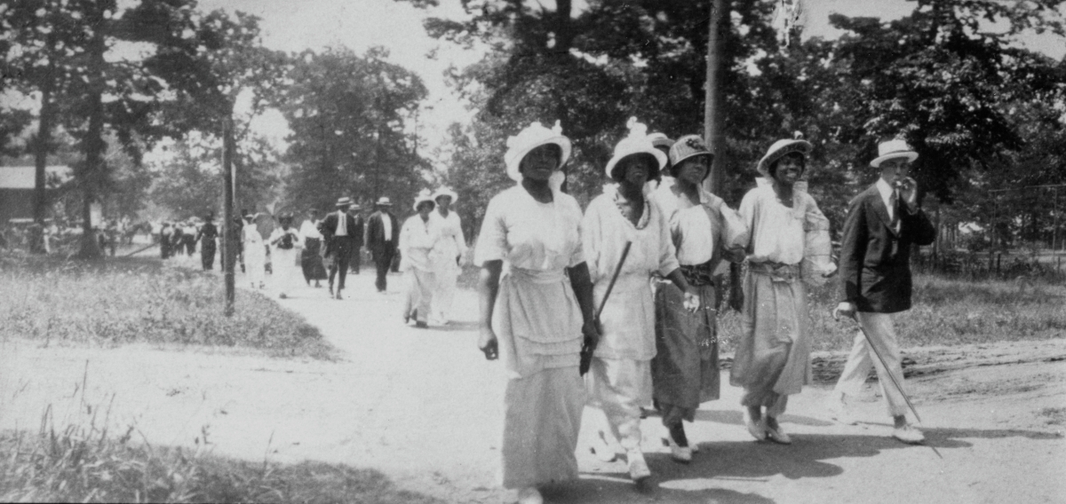 A black and white photo showing a four African American women and one man walking along a dirt lane. There are more individuals in small groups walking in the same direction on the path behind them. All are dressed in suits and dresses.