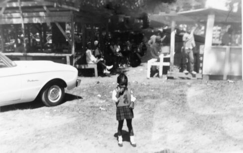 A young African American girl stands in front of wooden structures that other people are sitting or standing near in a park setting.