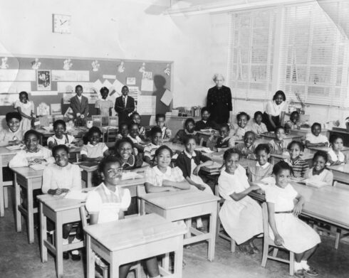 A black and white photo showing African American elementary school children sitting at desks and tables in their classroom for a class photo.