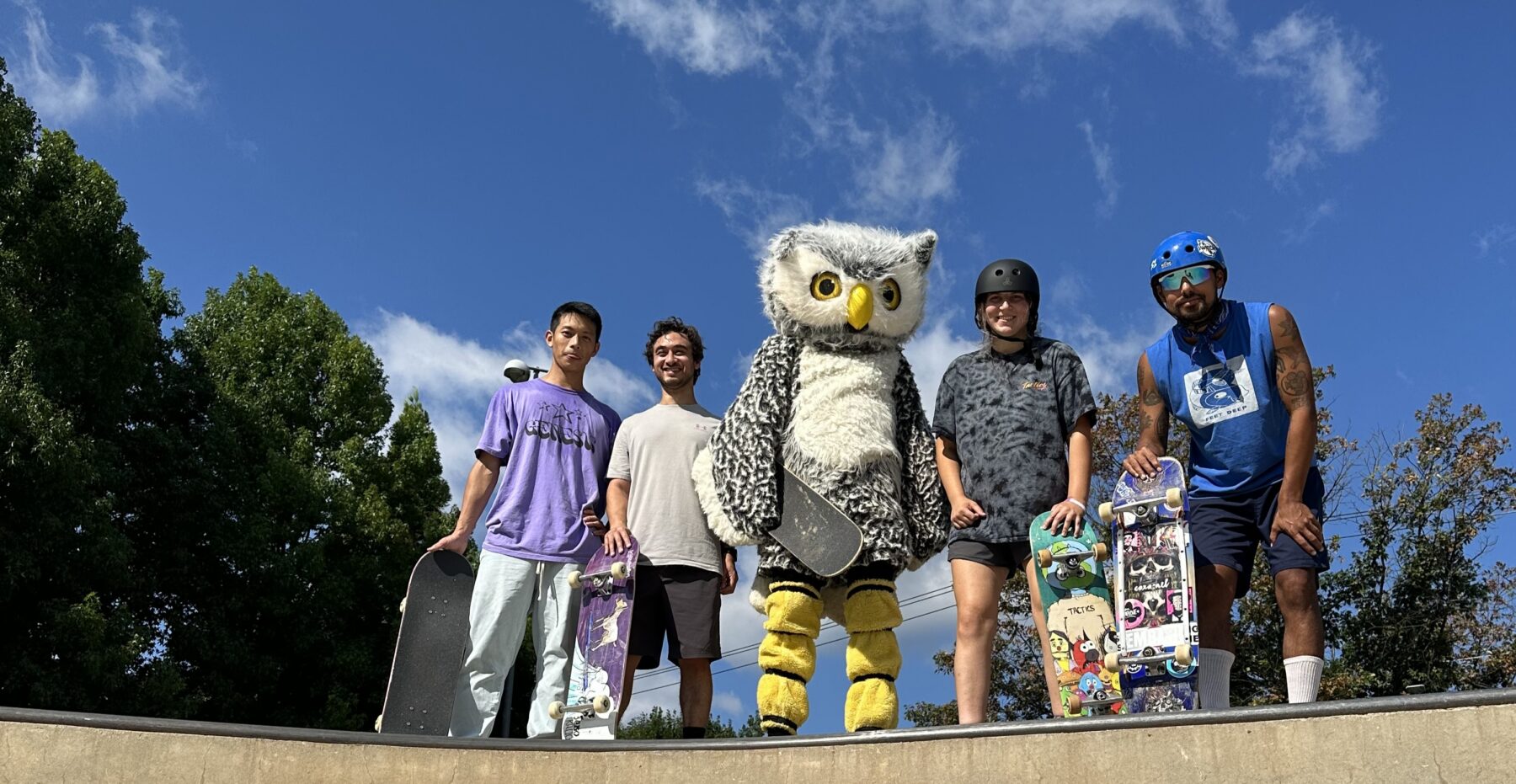 Otus the Owl with four other skateboarders holding skateboards and standing on a skateboarding ramp with a blue sky and trees in the background.