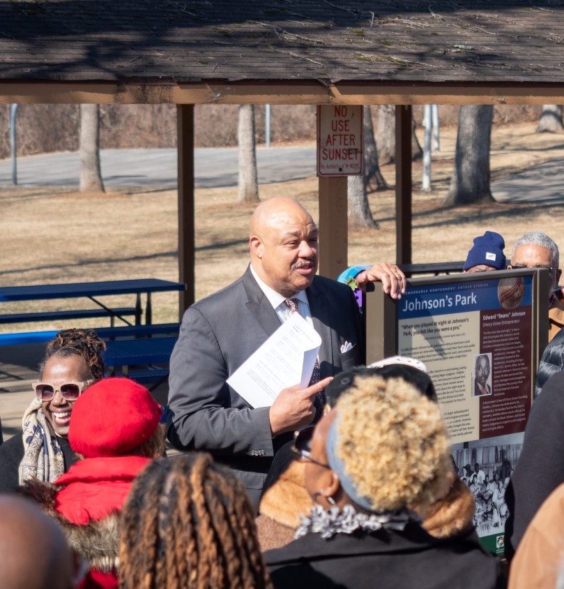 A crowd stands near an interpretive sign that reads "Johnson's Park"