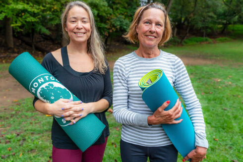 Two women pose holding yoga mats while standing in a park.