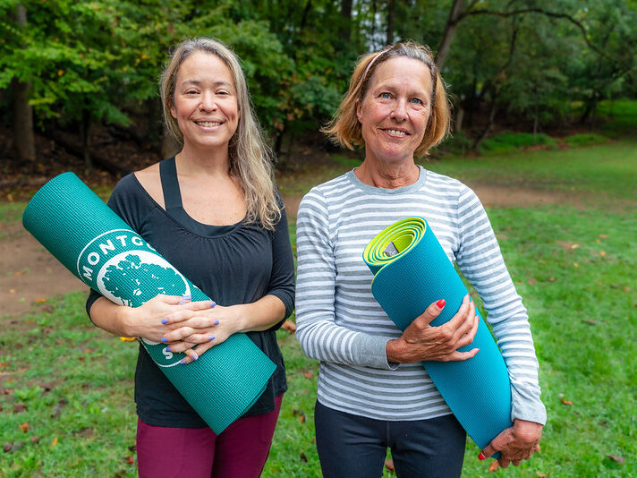 Two women pose holding yoga mats while standing in a park.