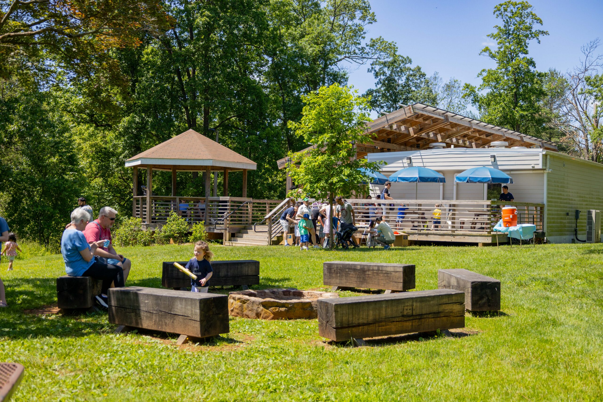 A fire pit with benches in a flat grass field. A patio and gazebo attached to a deck.