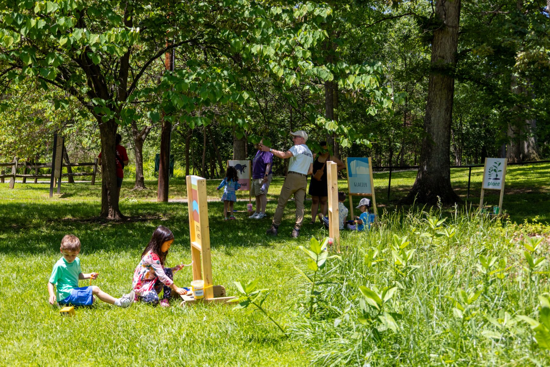 Children and adults standing and sitting in the grass near activity signs.