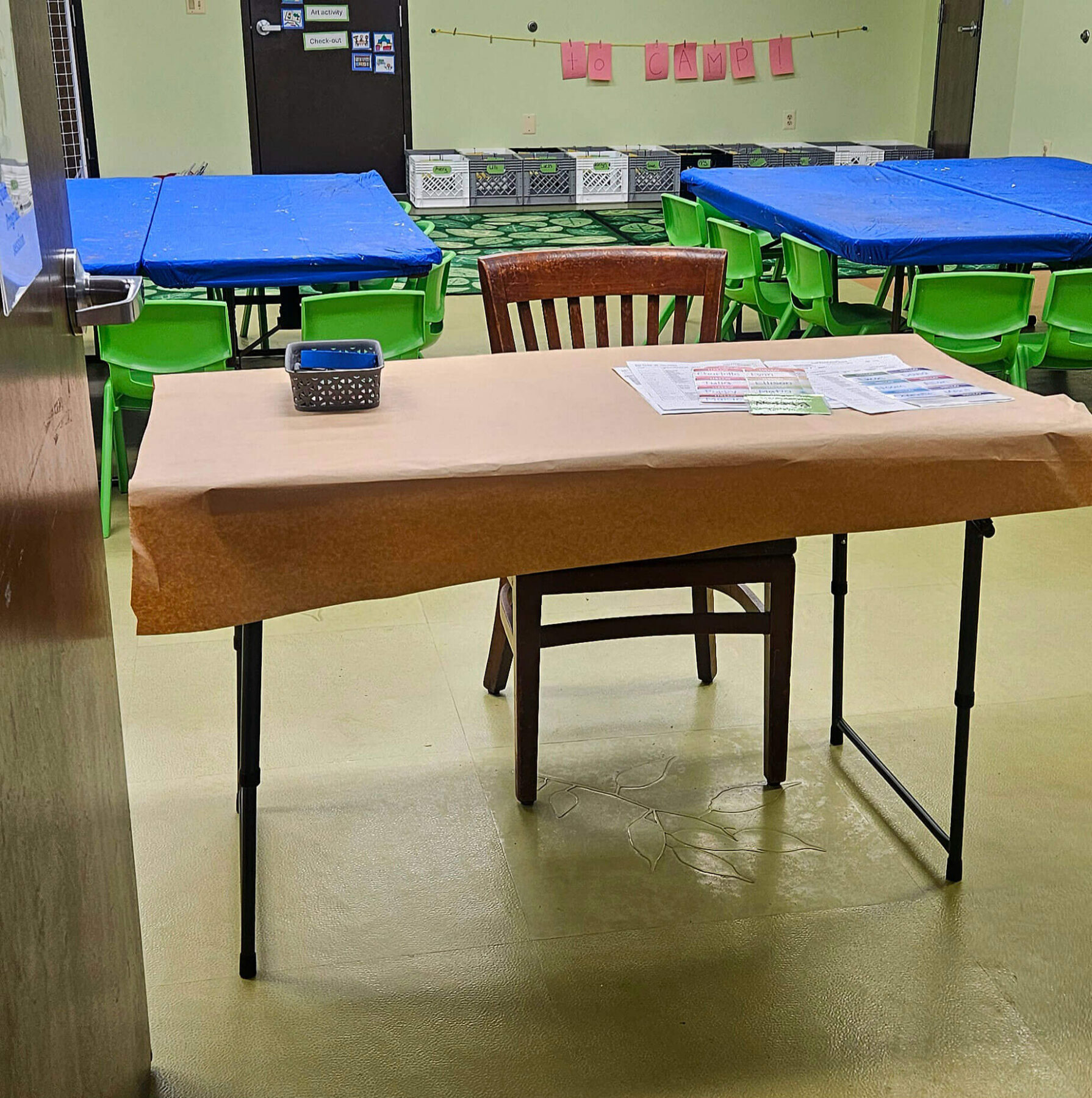 A table in the middle on the doorway just behind the doors to the auditorium with to longer tables in the background and empty crates on the floor behind the larger tables